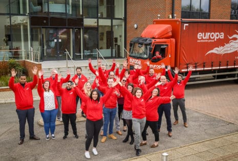 The Europa Road Heathrow team are stood, cheering outside Europa Heathrow in red, branded hoodies.